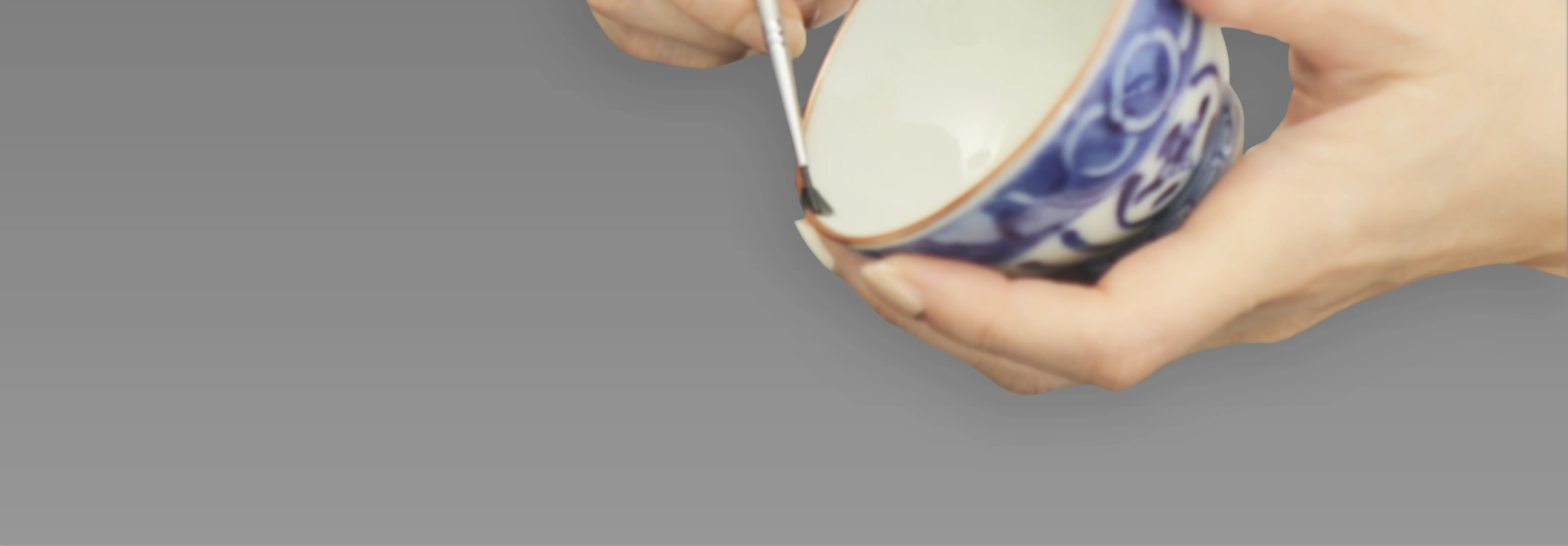 A brush applying lacquer to the rim of a blue-and-white bowl held in both hands.
